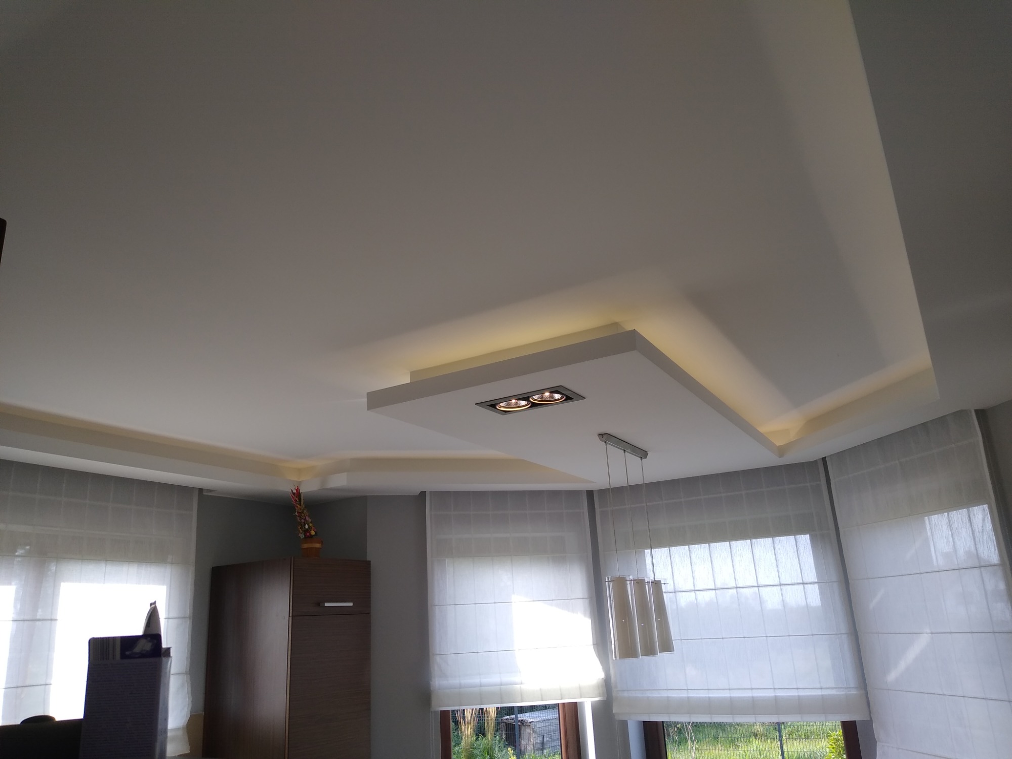 Jasne wnętrze z podwieszanym sufitem, oświetleniem punktowym i dekoracyjnym, widoczne rolety rzymskie w oknach.