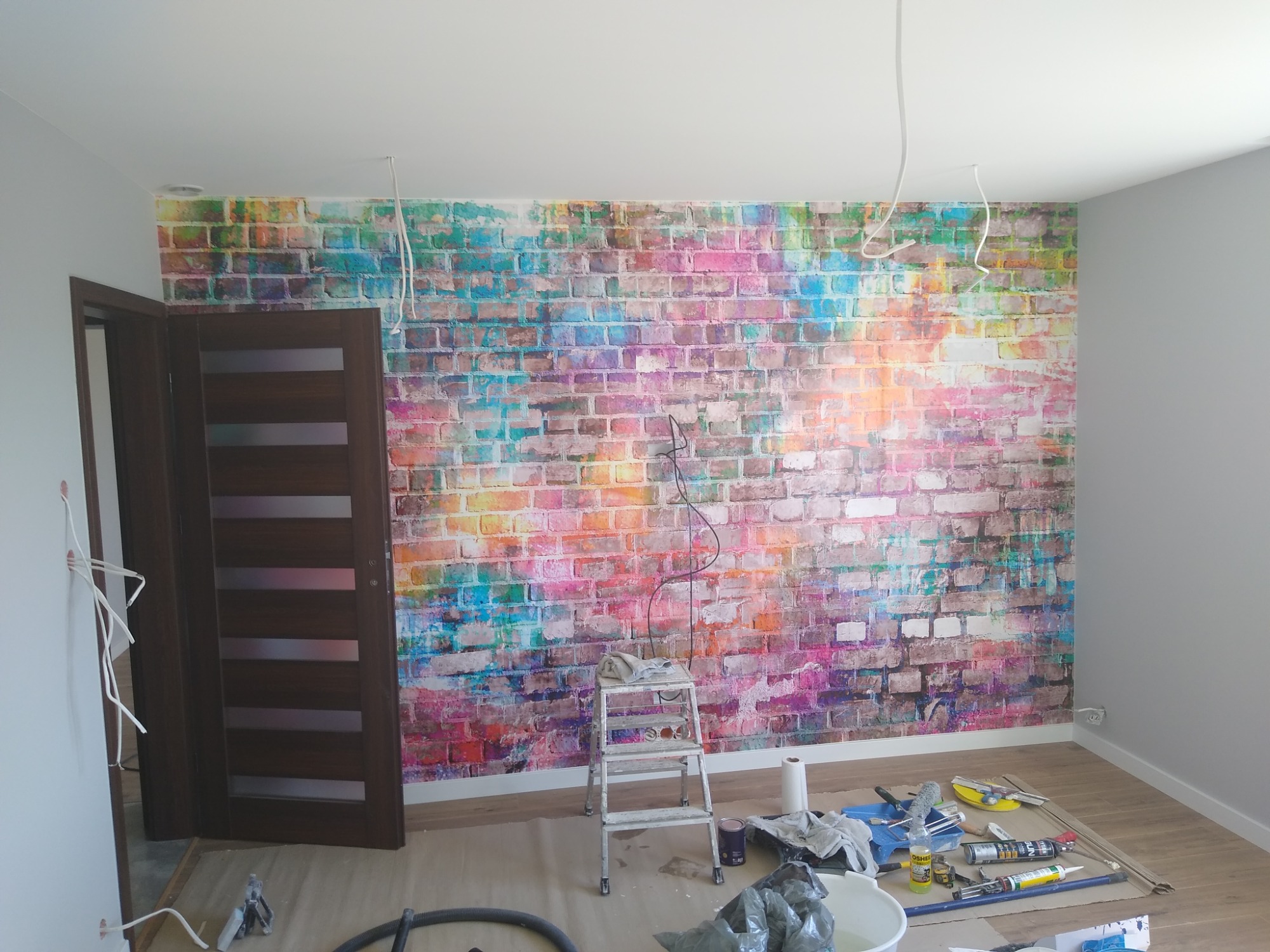 Pokój w trakcie dekorowania: ściana z kolorową tapetą imitującą cegły, drabina, narzędzia malarskie i przewody wystające ze ściany.