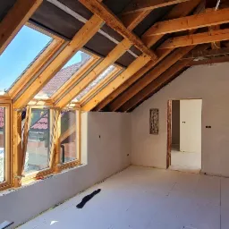 Poddasze po nałożeniu szarego tynku maszynowego, widoczne drewniane krokwie, okna dachowe i przygotowane podłoże pod podłogę.