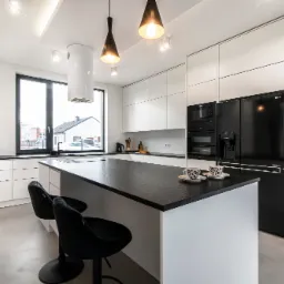 Nowoczesna kuchnia w bieli i czerni z wyspą, wyposażona w minimalistyczne szafki, czarny sprzęt AGD i dwie czarne, welurowe hokery przy blacie.
