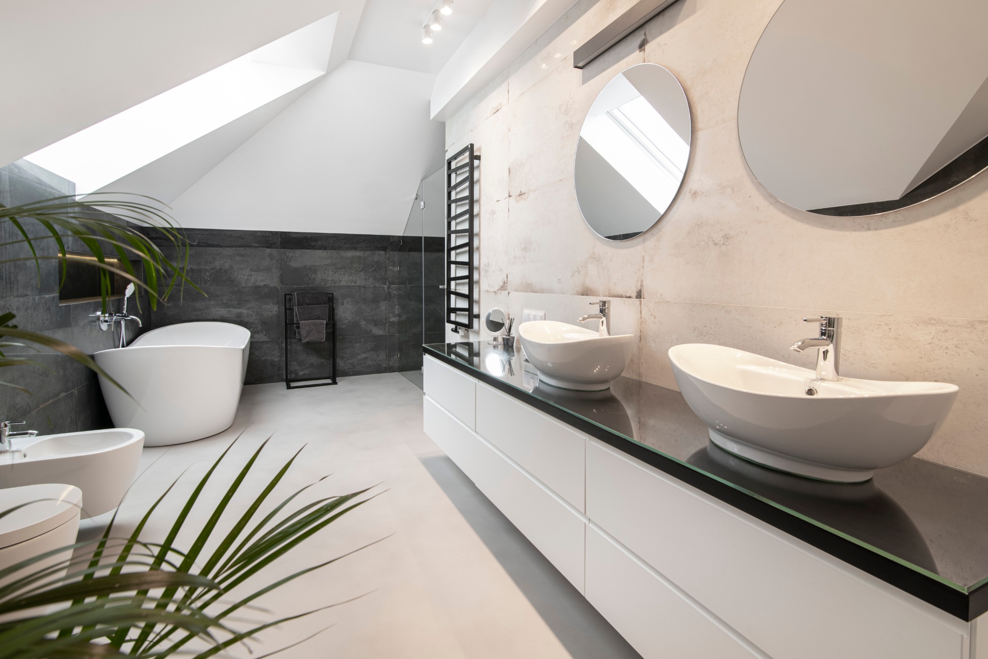 Nowoczesna łazienka na poddaszu z białą wolnostojącą wanną, bidetem, dwiema owalnymi umywalkami na czarnym blacie i okrągłymi lustrami, oświetlona światłem z okna dachowego.