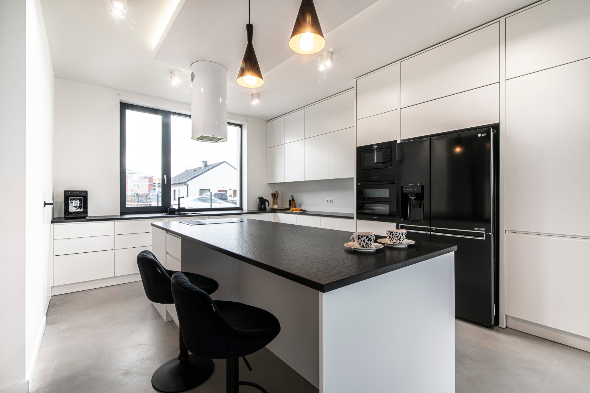 Nowoczesna kuchnia w bieli i czerni z wyspą, wyposażona w minimalistyczne szafki, czarny sprzęt AGD i dwie czarne, welurowe hokery przy blacie.