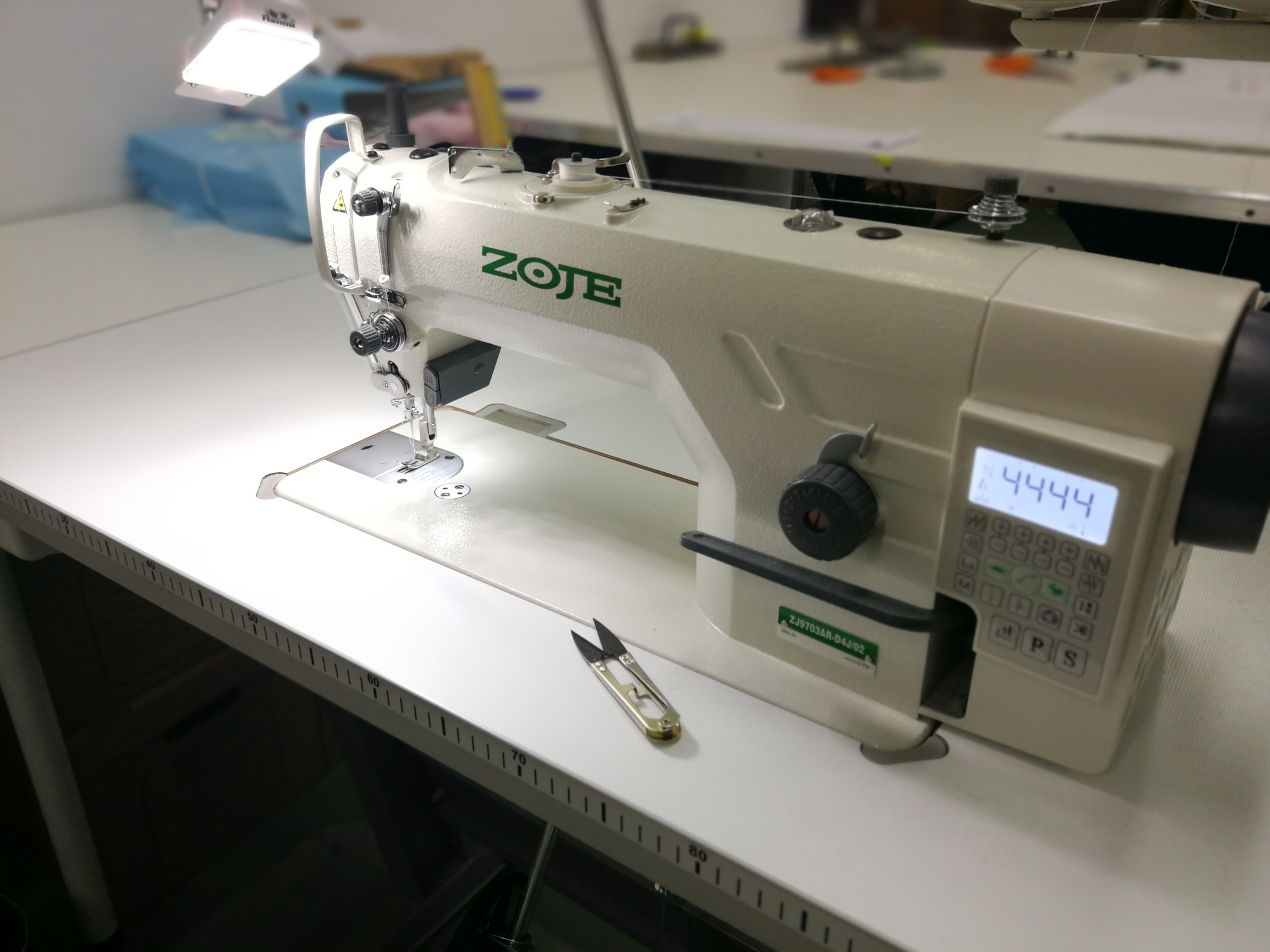 Profesjonalna maszyna do szycia marki ZOJE na białym stole z miarką, obok leżą nożyczki do nici.