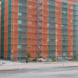 Wielopiętrowy budynek w trakcie renowacji, osłonięty siatką ochronną i rusztowaniami, z widoczną metalową klatką schodową na elewacji i pracownikiem na dachu.