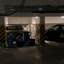 Ozon Piorun - Ozonowanie Gdańsk, Trójmiasto, Cała Polska - Pracownik w kombinezonie stoi w garażu podziemnym obok sprzętu do ozonowania, widoczne dwa samochody z naklejkami ostrzegawczymi.