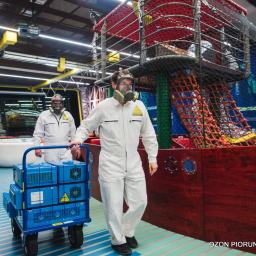 Ozon Piorun - Ozonowanie Gdańsk, Trójmiasto, Cała Polska - Dwaj pracownicy w kombinezonach i maskach gazowych, jeden ciągnie wózek z niebieskimi generatorami ozonu wewnątrz sali zabaw z konstrukcją przypominającą statek piracki i labirynt linowy.