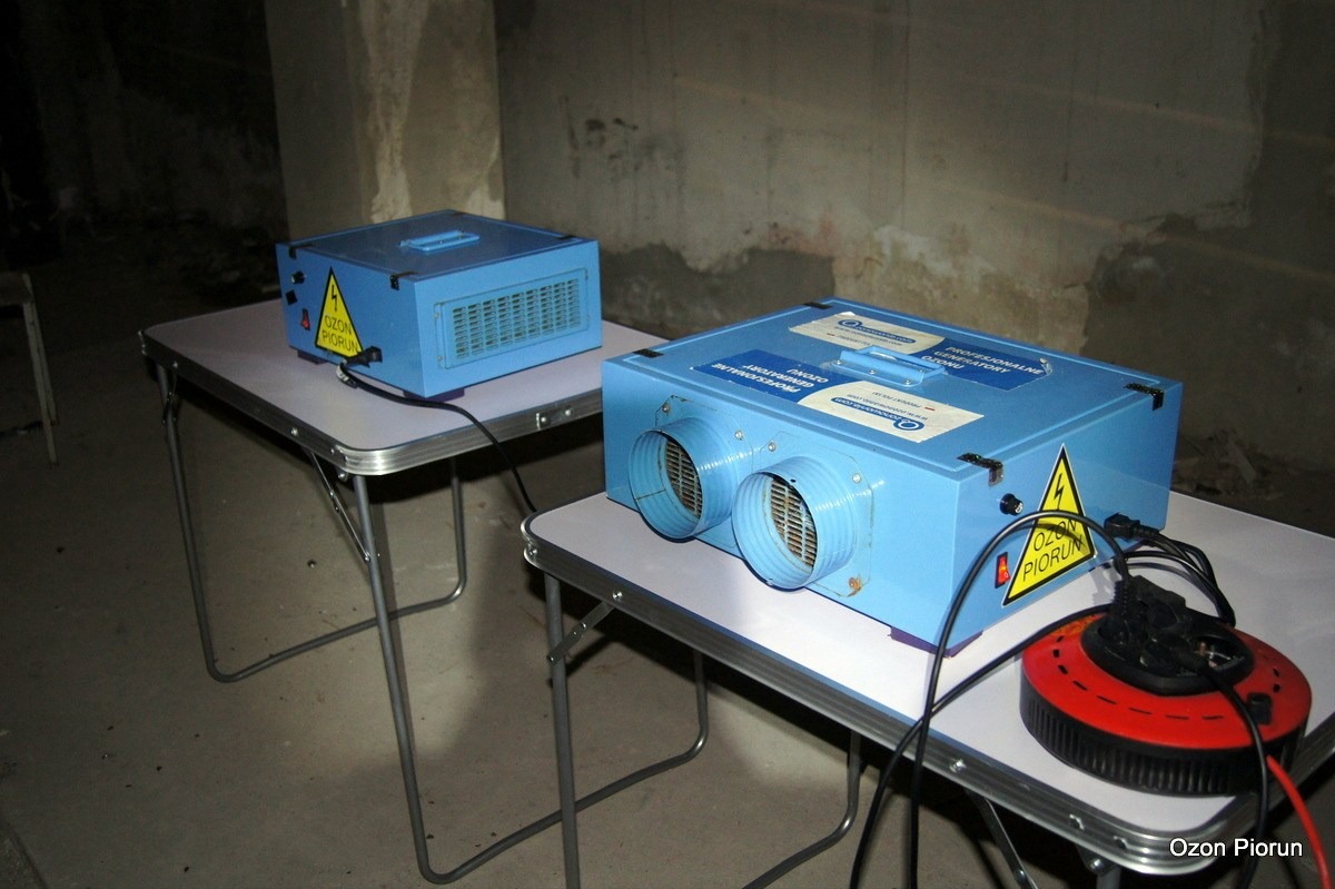 Dwa niebieskie generatory ozonu na składanych stołach z aluminiowymi nogami, w surowym pomieszczeniu z betonowymi ścianami. Jeden generator ma dwa okrągłe wyloty powietrza z kratkami.