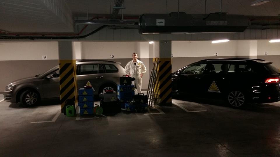 Pracownik w kombinezonie stoi w garażu podziemnym obok sprzętu do ozonowania, widoczne dwa samochody z naklejkami ostrzegawczymi.