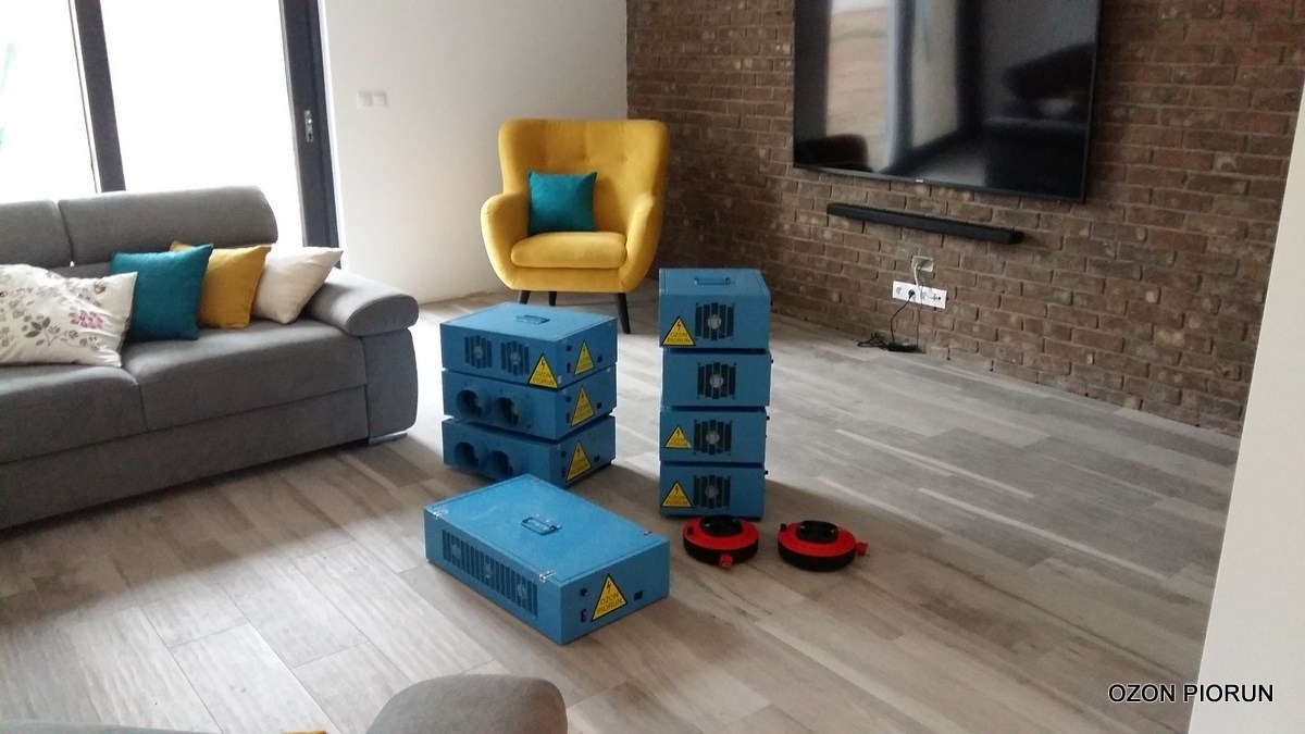 Niebieskie generatory ozonu ustawione w nowoczesnym salonie z szarą sofą, żółtym fotelem i ceglaną ścianą z telewizorem.