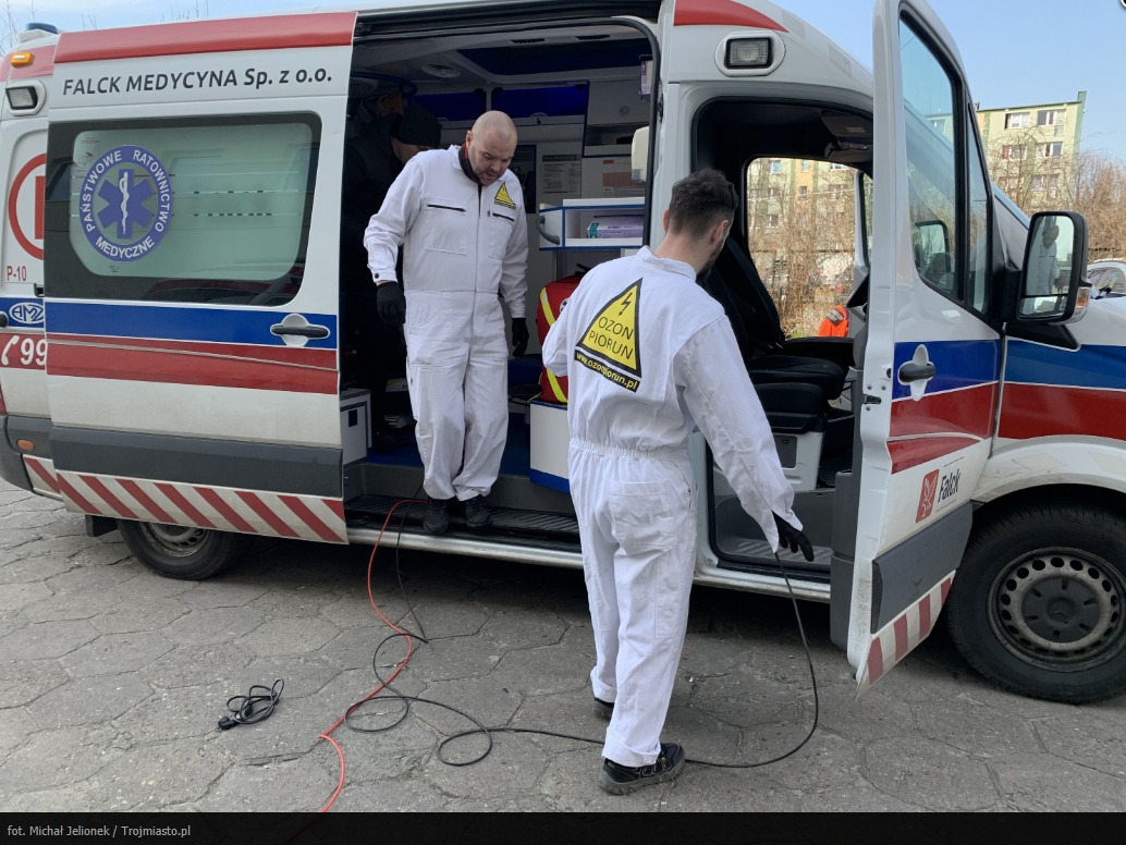 Dwóch techników w białych kombinezonach przy ambulansie Falck Medycyna, jeden wchodzi do środka, drugi podłącza przewód urządzenia do ozonowania z logo 'Ozon Piorun' na plecach.