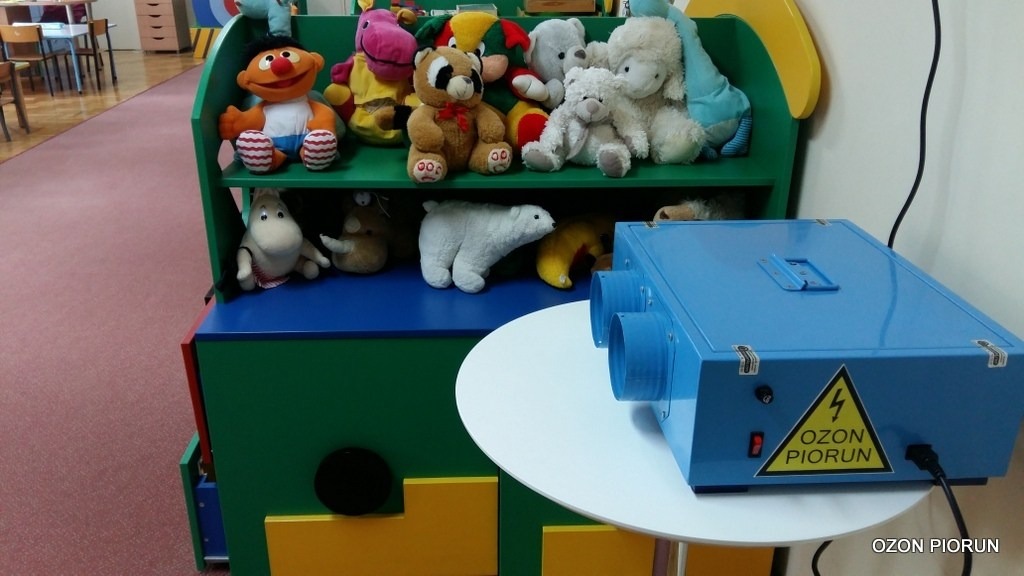 Niebieskie urządzenie do ozonowania, marki Ozon Piorun, ustawione na białym stoliku w pokoju dziecięcym, na tle regału z pluszakami.