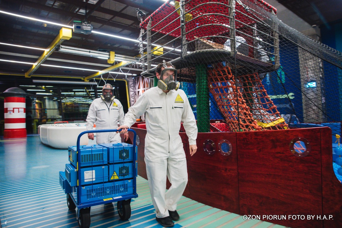 Dwaj pracownicy w kombinezonach i maskach gazowych, jeden ciągnie wózek z niebieskimi generatorami ozonu wewnątrz sali zabaw z konstrukcją przypominającą statek piracki i labirynt linowy.