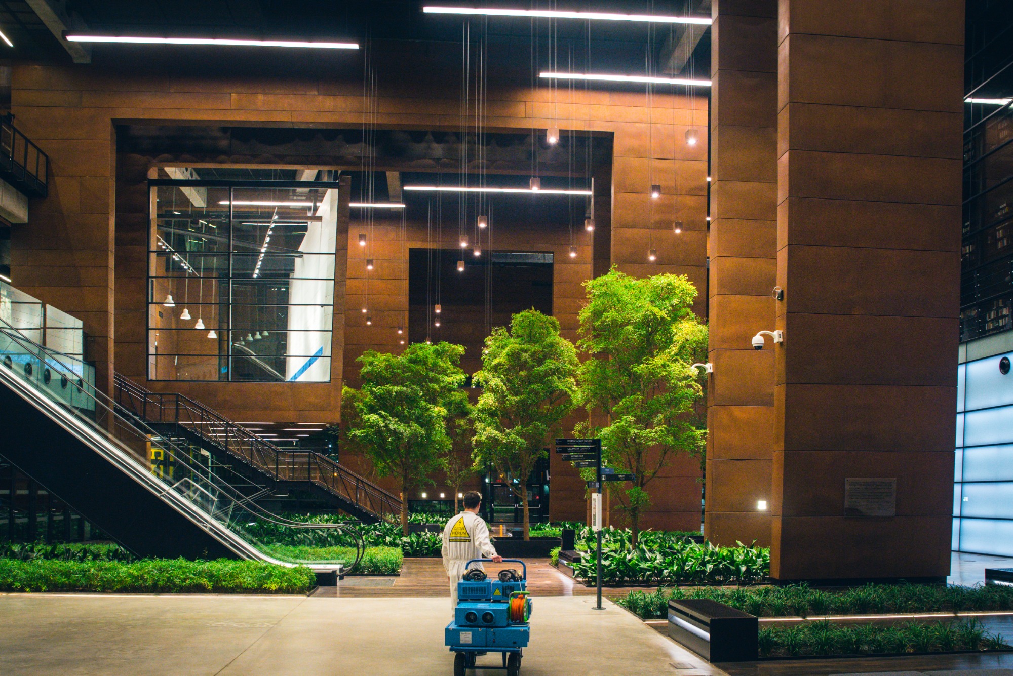 Pracownik w kombinezonie z logo Ozon Piorun ciągnie niebieski wózek z urządzeniem do ozonowania w przestronnym, nowoczesnym wnętrzu z roślinami, schodami ruchomymi i oświetleniem punktowym.