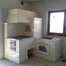 Nowo wybudowany, kaflowy piec kuchenny z piekarnikiem, płytą grzewczą i okapem w surowym wnętrzu. Widoczne niedokończone ściany i okno.