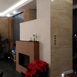 Nowoczesny salon z obudowanym kominkiem w odcieniach beżu i brązu, podświetlany sufit, dekoracje na kominku i czerwona poinsecja w doniczce.