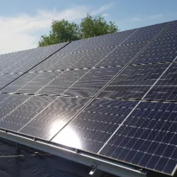 Instalacja fotowoltaiczna z widocznymi panelami słonecznymi na konstrukcji wsporczej, w tle błękitne niebo z chmurami i zieleń drzew.