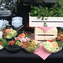 Bufet sałatkowy z różnorodnymi składnikami w szklanych misach, udekorowany ziołami w drewnianej skrzynce i czerwoną kratą, na czarnym stole, z podgrzewaczami w tle.