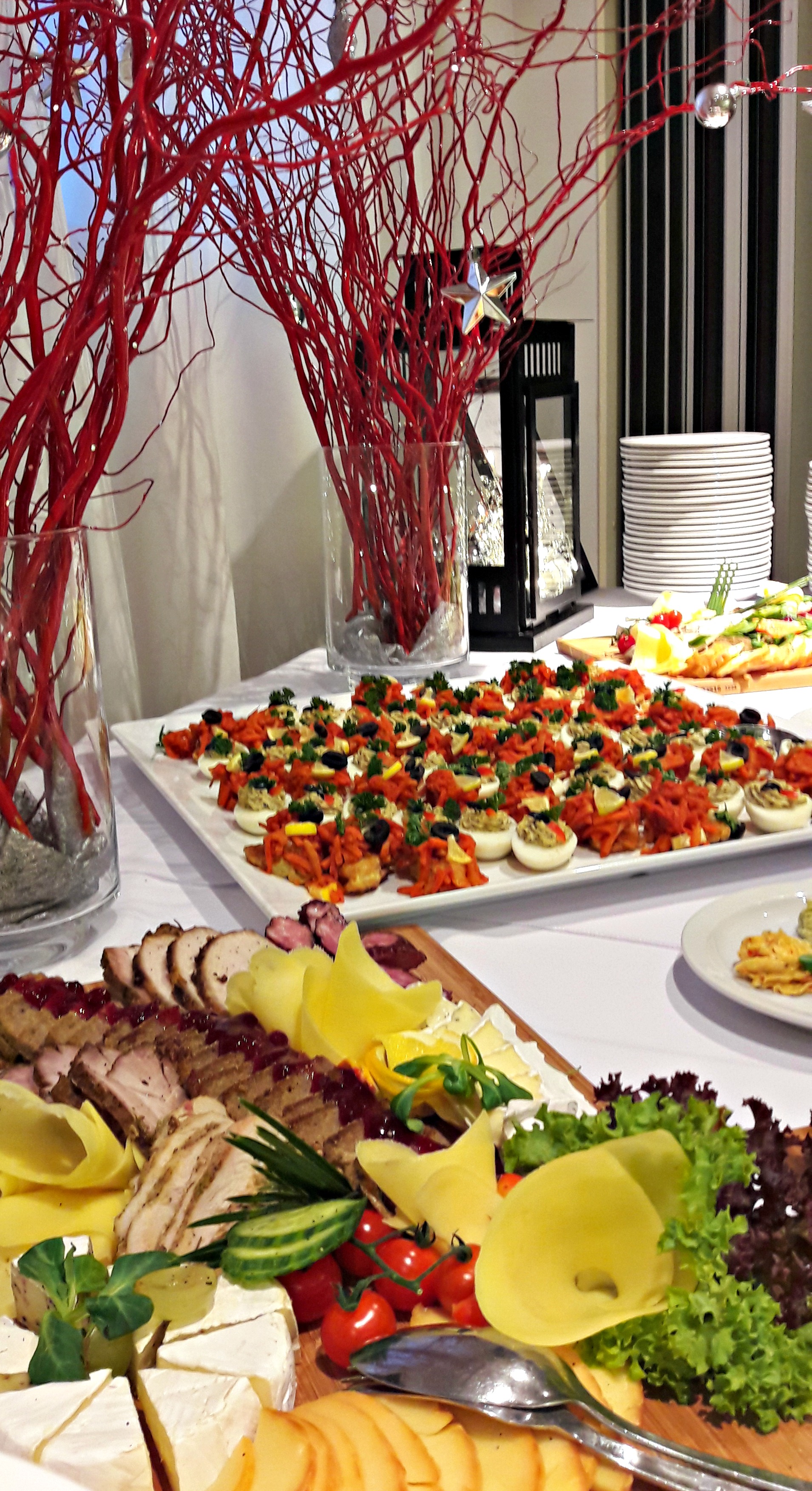 Dekoracyjny stół cateringowy z jajkami faszerowanymi, wędlinami, serami i warzywami, ozdobiony czerwonymi gałązkami w wazonach.