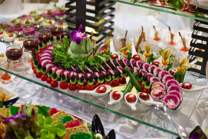 Dekoracyjnie ułożone przekąski na szklanym stole: mini szaszłyki z pomidorków i oliwek, rolki z buraka, sałatki w szklankach, papryczki nadziewane na łyżeczkach, całość udekorowana karczochem...