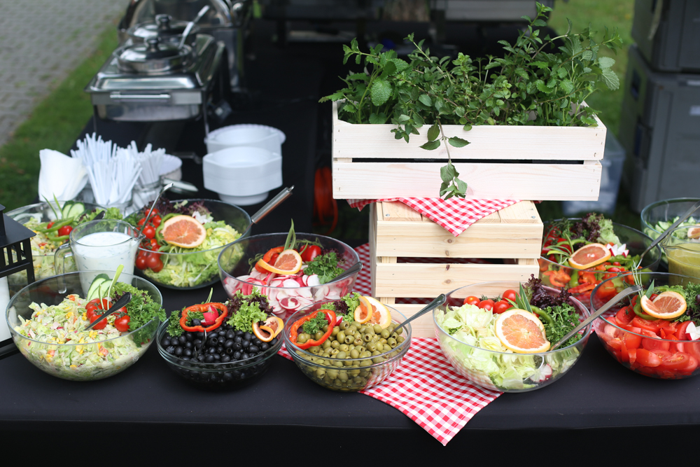 Bufet sałatkowy z różnorodnymi składnikami w szklanych misach, udekorowany ziołami w drewnianej skrzynce i czerwoną kratą, na czarnym stole, z podgrzewaczami w tle.