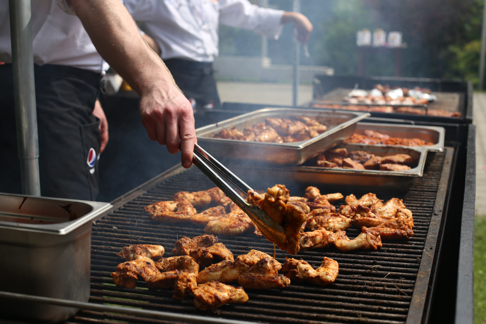 Przygotowywanie grillowanych skrzydełek z przyprawami na ruszcie przez osobę w fartuchu, widoczne także metalowe pojemniki z mięsem i para unosząca się znad grilla.