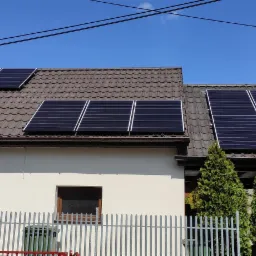 Panele słoneczne zamontowane na brązowym dachu domu jednorodzinnego z jasną elewacją, widoczne trzy rzędy paneli, ogrodzenie metalowe i drzewka iglaste.