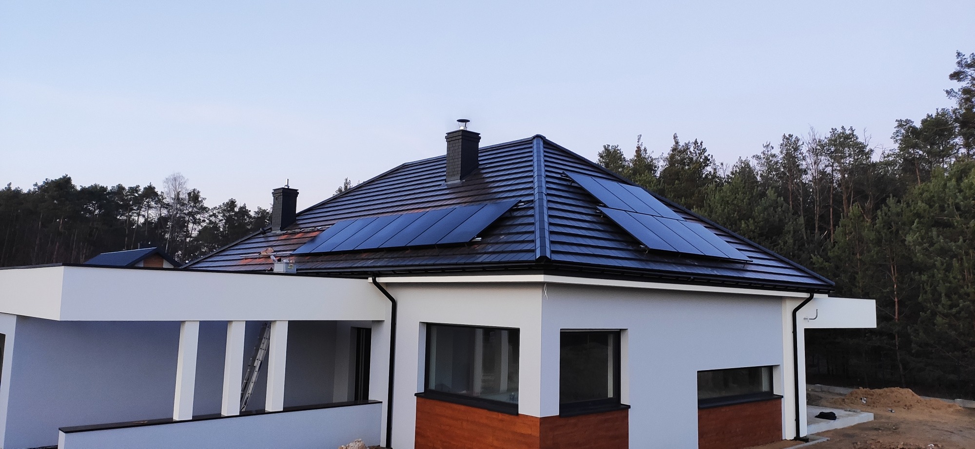 Nowoczesny dom jednorodzinny z ciemną dachówką i panelami słonecznymi na dachu, widok z boku, w tle las.