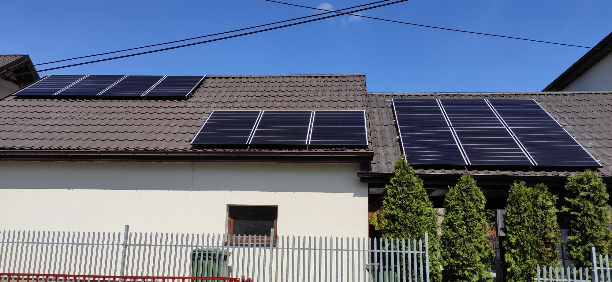 Panele słoneczne zamontowane na brązowym dachu domu jednorodzinnego z jasną elewacją, widoczne trzy rzędy paneli, ogrodzenie metalowe i drzewka iglaste.