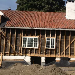 Wznoszenie drewnianej konstrukcji domu z widoczną więźbą, oknami z białymi ramami i czerwoną dachówką, z przygotowanym terenem wokół.