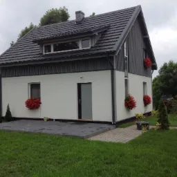 Nowoczesny dom z dwuspadowym dachem pokrytym ciemną dachówką, białą elewacją, szarymi elementami wykończeniowymi i czerwonymi kwiatami w oknach, otoczony zielonym trawnikiem i drzewami.