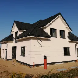 Nowoczesny dom jednorodzinny z białą elewacją i czarnym dachem, widoczny na etapie wykończenia, z elementami instalacji zewnętrznej i drabina przy wejściu.