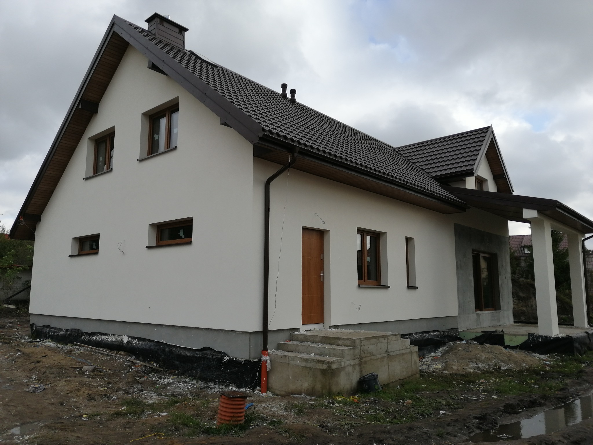 Nowo otynkowany dom jednorodzinny z brązowym dachem, widoczne niedokończone schody wejściowe i przygotowany teren wokół budynku.