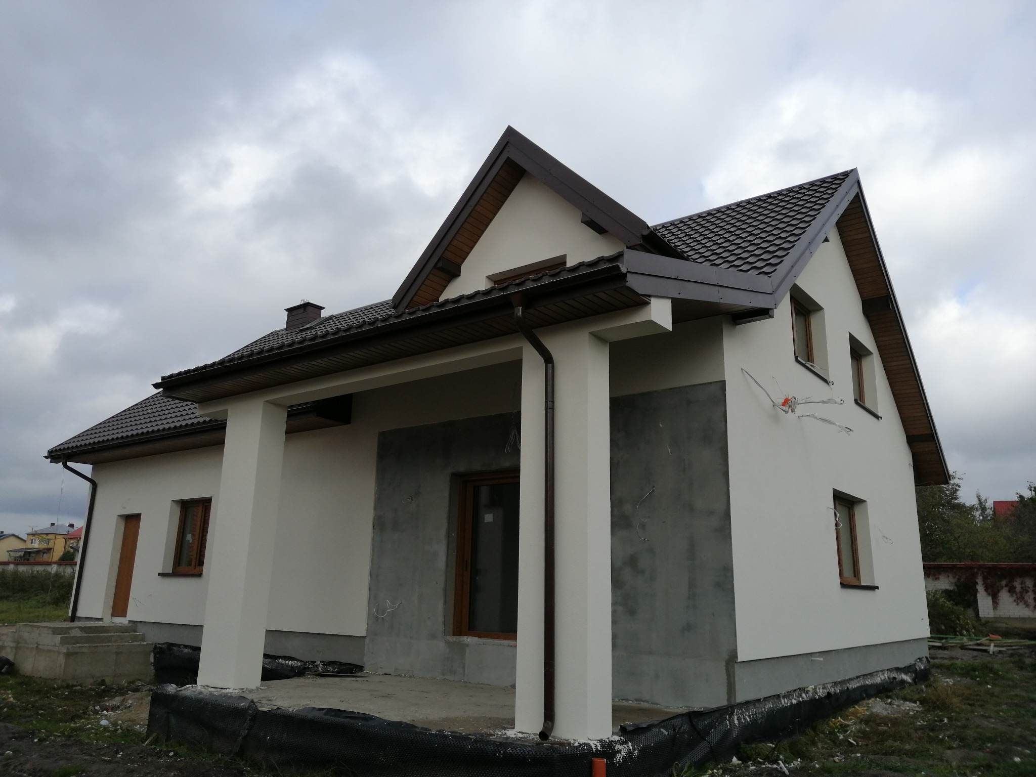 Dom jednorodzinny w trakcie prac elewacyjnych: częściowo otynkowana ściana, widoczne przewody elektryczne, brązowa dachówka i stolarka okienna.