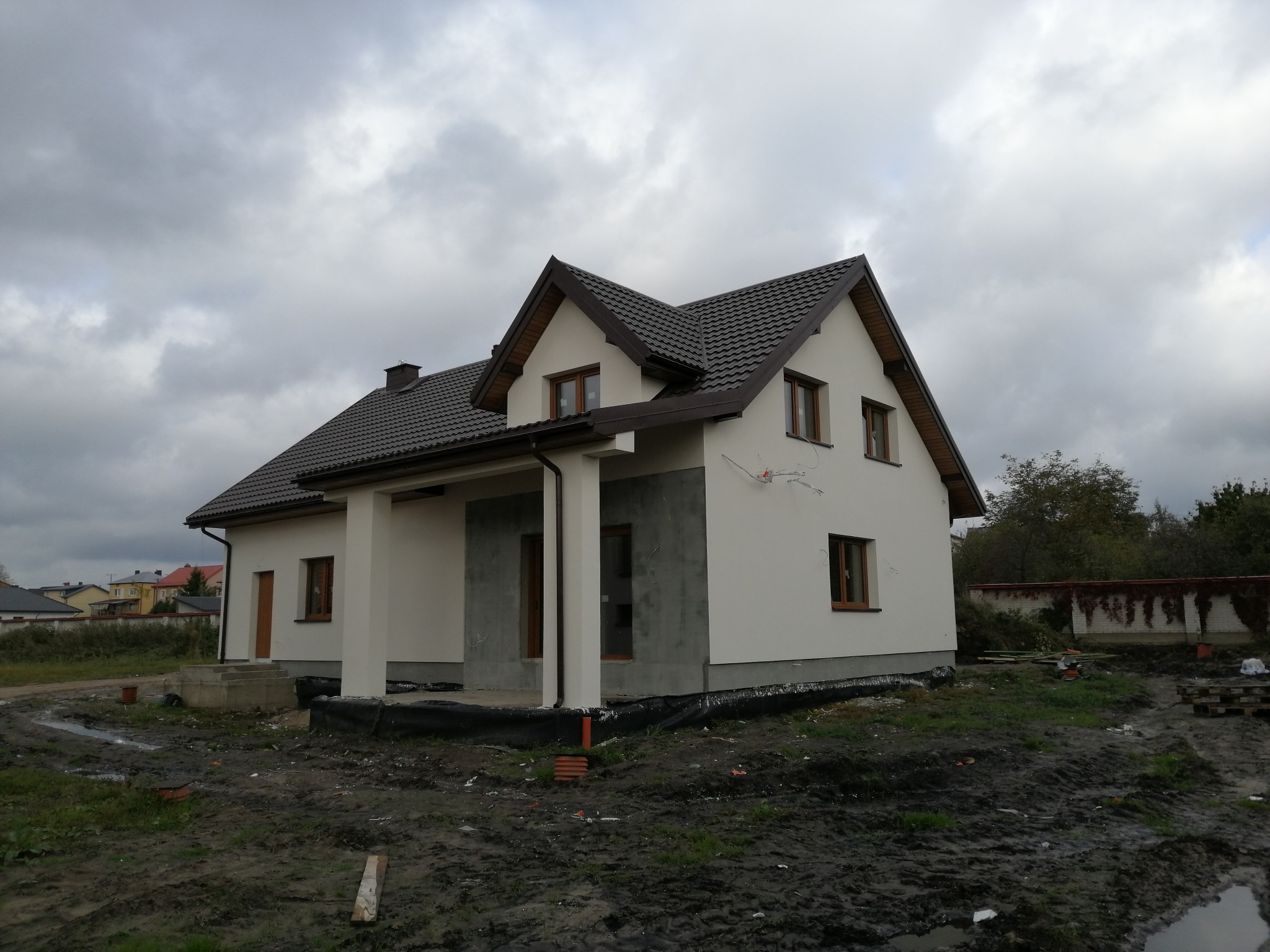 Dom jednorodzinny z grafitową dachówką i jasną elewacją w trakcie wykańczania, widoczne fragmenty szarego ocieplenia przy fundamentach, teren wokół domu częściowo błotnisty.