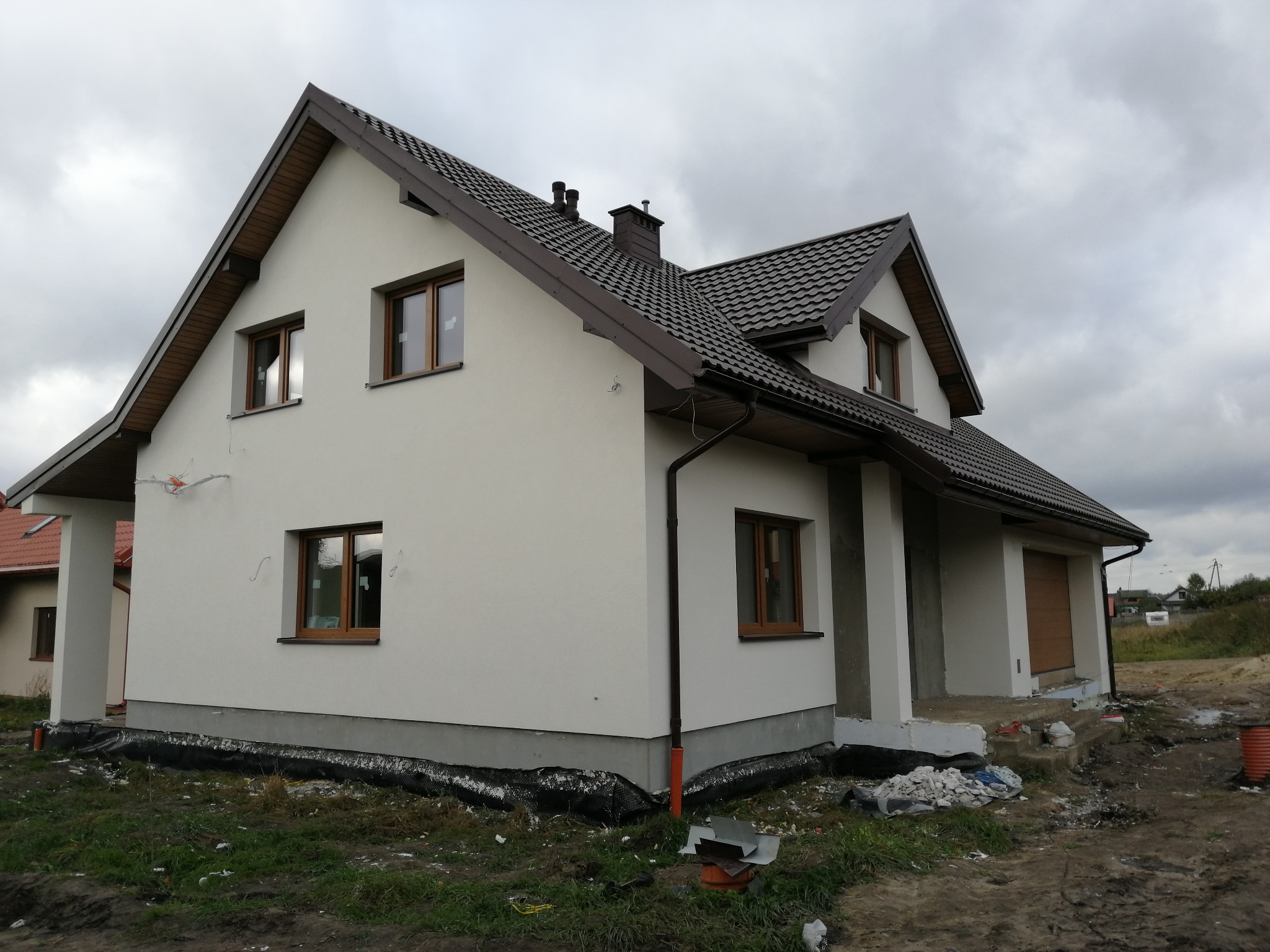 Nowo wybudowany dom jednorodzinny z jasną elewacją, brązowym dachem i drewnianą stolarką okienną, widoczne pozostałości materiałów budowlanych wokół budynku.
