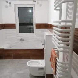 Nowoczesna łazienka z wanną obudowaną drewnopodobnymi płytkami, wiszącą toaletą, grzejnikiem drabinkowym z pomarańczowym ręcznikiem i szarą podłogą, oświetlona trzema punktowymi lampami.