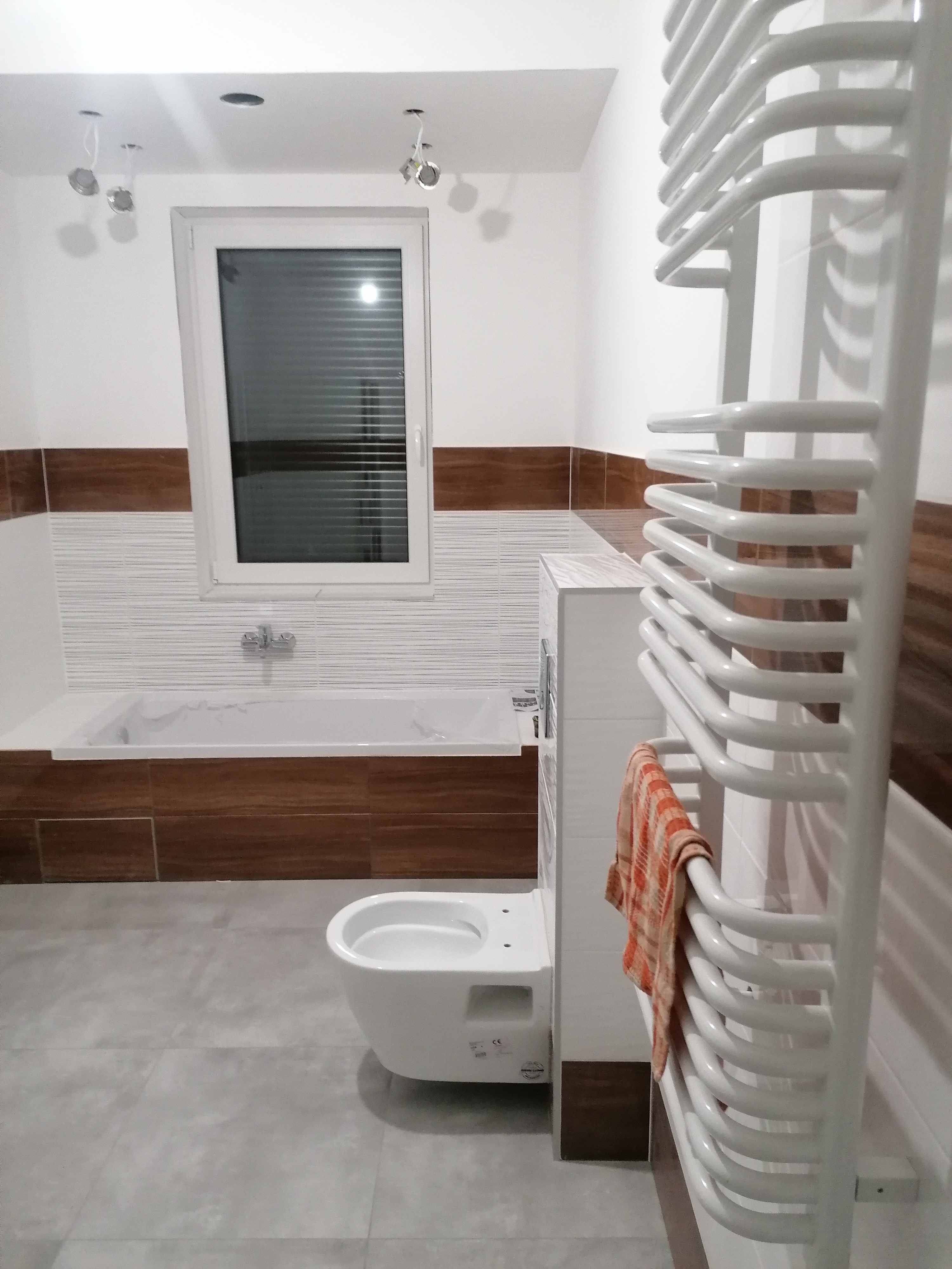 Nowoczesna łazienka z wanną obudowaną drewnopodobnymi płytkami, wiszącą toaletą, grzejnikiem drabinkowym z pomarańczowym ręcznikiem i szarą podłogą, oświetlona trzema punktowymi lampami.