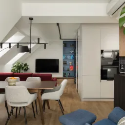 Nowoczesne wnętrze mieszkania na poddaszu: stół z krzesłami, czerwona sofa, zabudowa kuchenna z drewnianymi akcentami i niebieski regał z książkami.