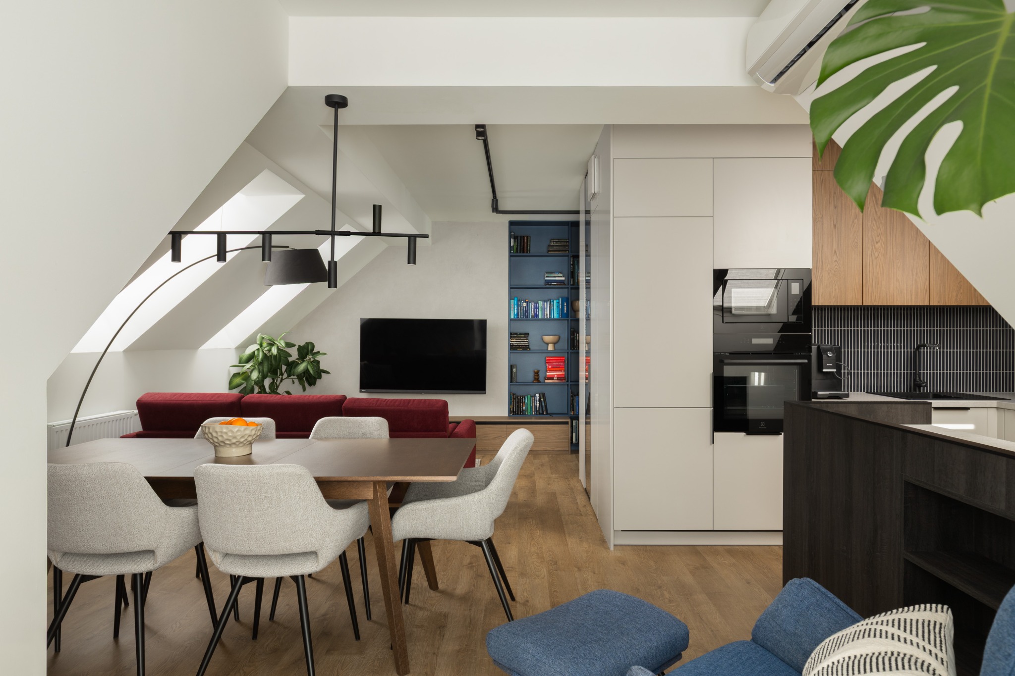 Nowoczesne wnętrze mieszkania na poddaszu: stół z krzesłami, czerwona sofa, zabudowa kuchenna z drewnianymi akcentami i niebieski regał z książkami.