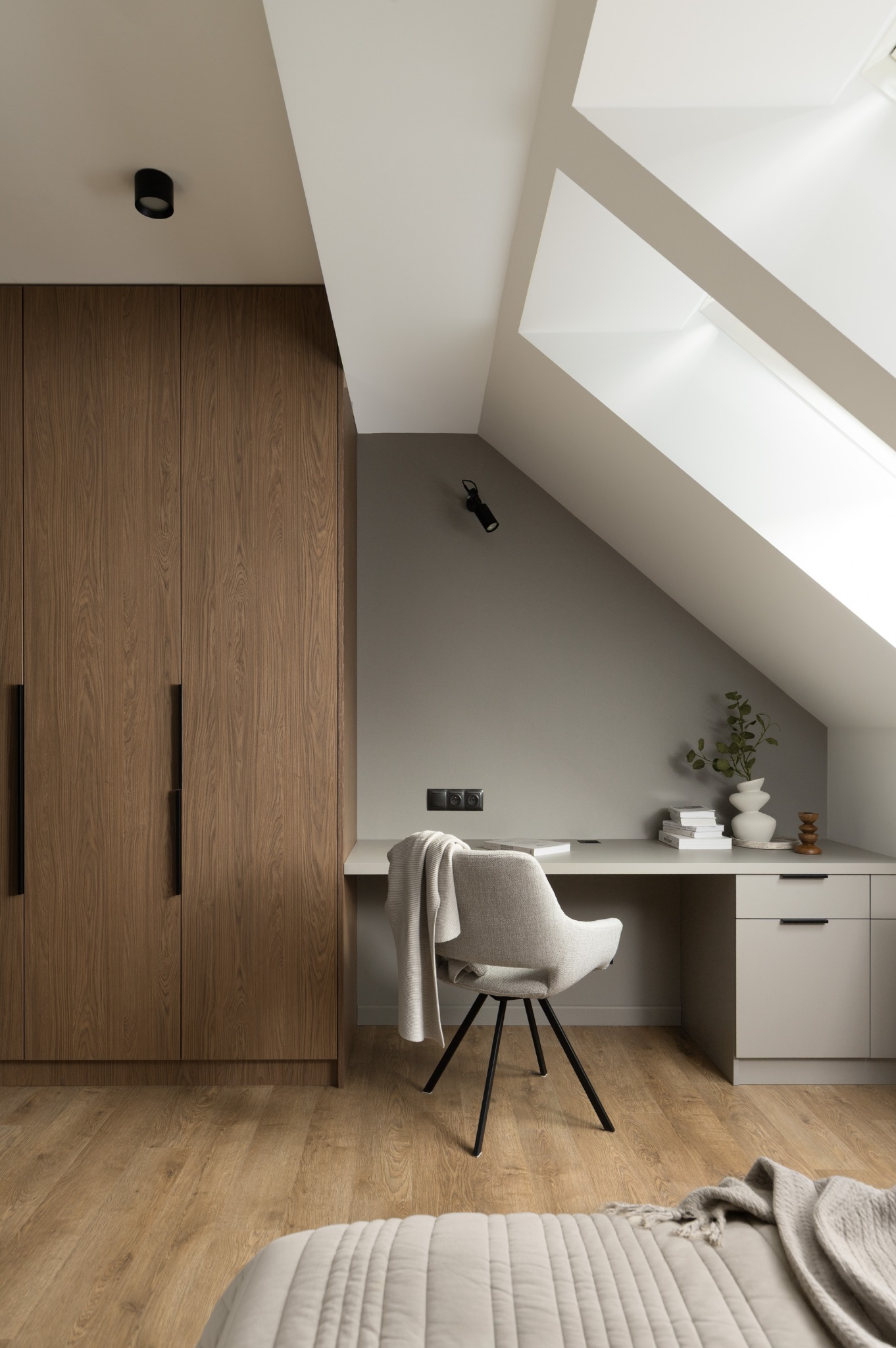 Pokój na poddaszu ze skośnym oknem dachowym, drewnianą szafą i biurkiem z krzesłem. Neutralna kolorystyka, minimalistyczny design.