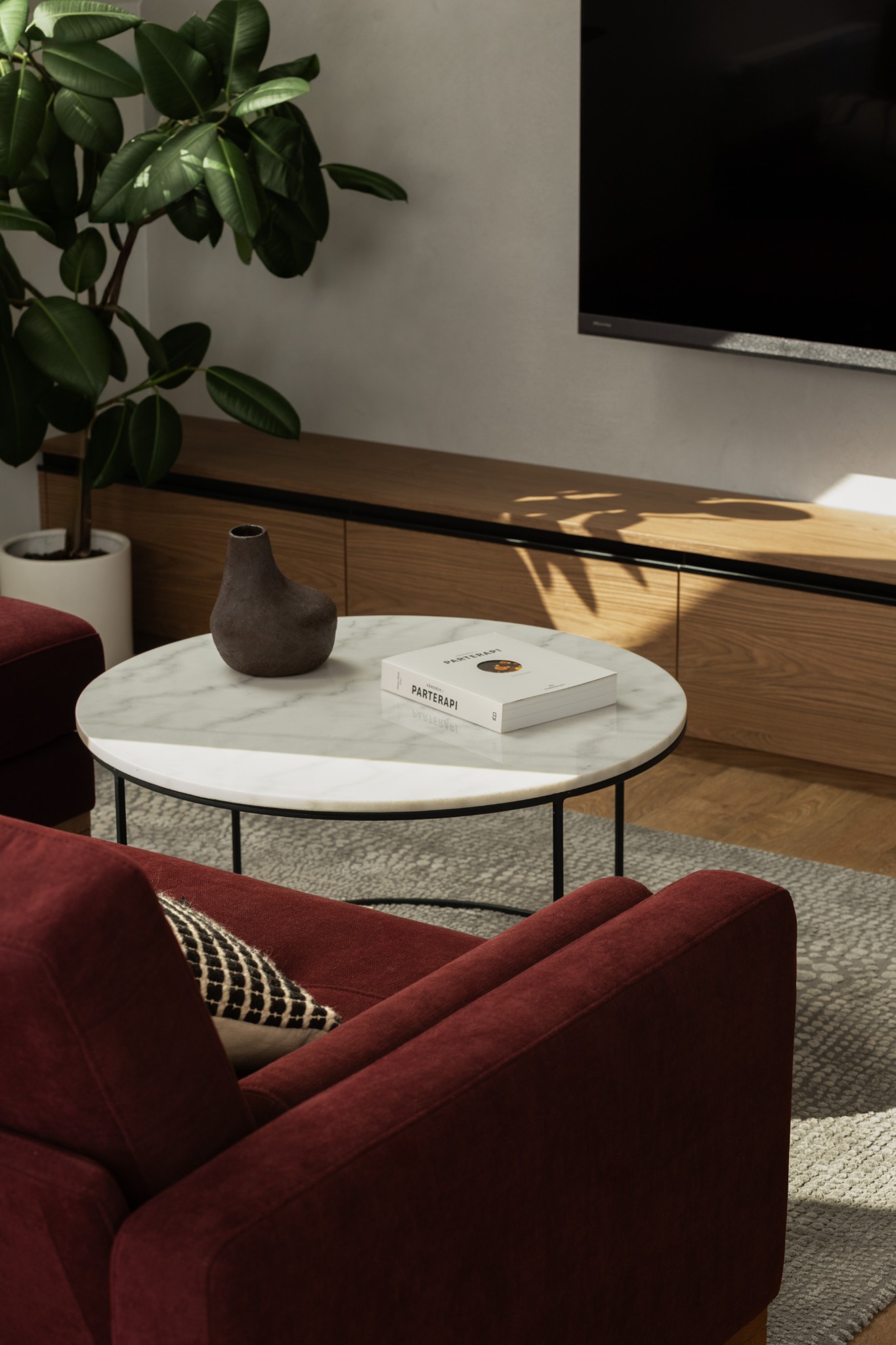 Nowoczesny salon z burgundową sofą, stolikiem z marmurowym blatem, wazonem i książką. W tle szafka RTV i telewizor, doświetlone naturalnym światłem.