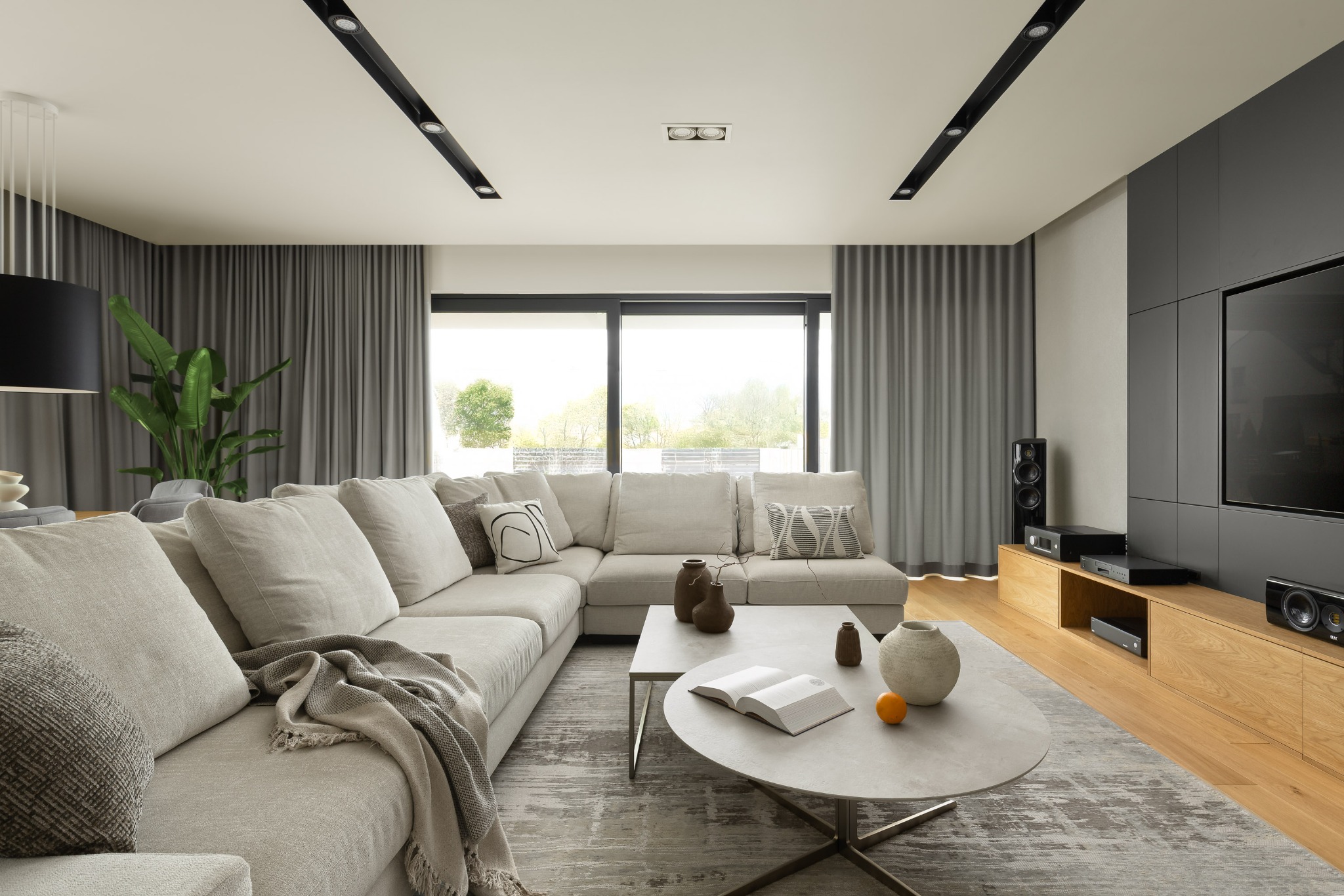 Jasny salon z narożną sofą, okrągłym stolikiem kawowym i zabudową TV. Stonowana kolorystyka, duże okno z widokiem na zieleń, minimalistyczne oświetlenie sufitowe.