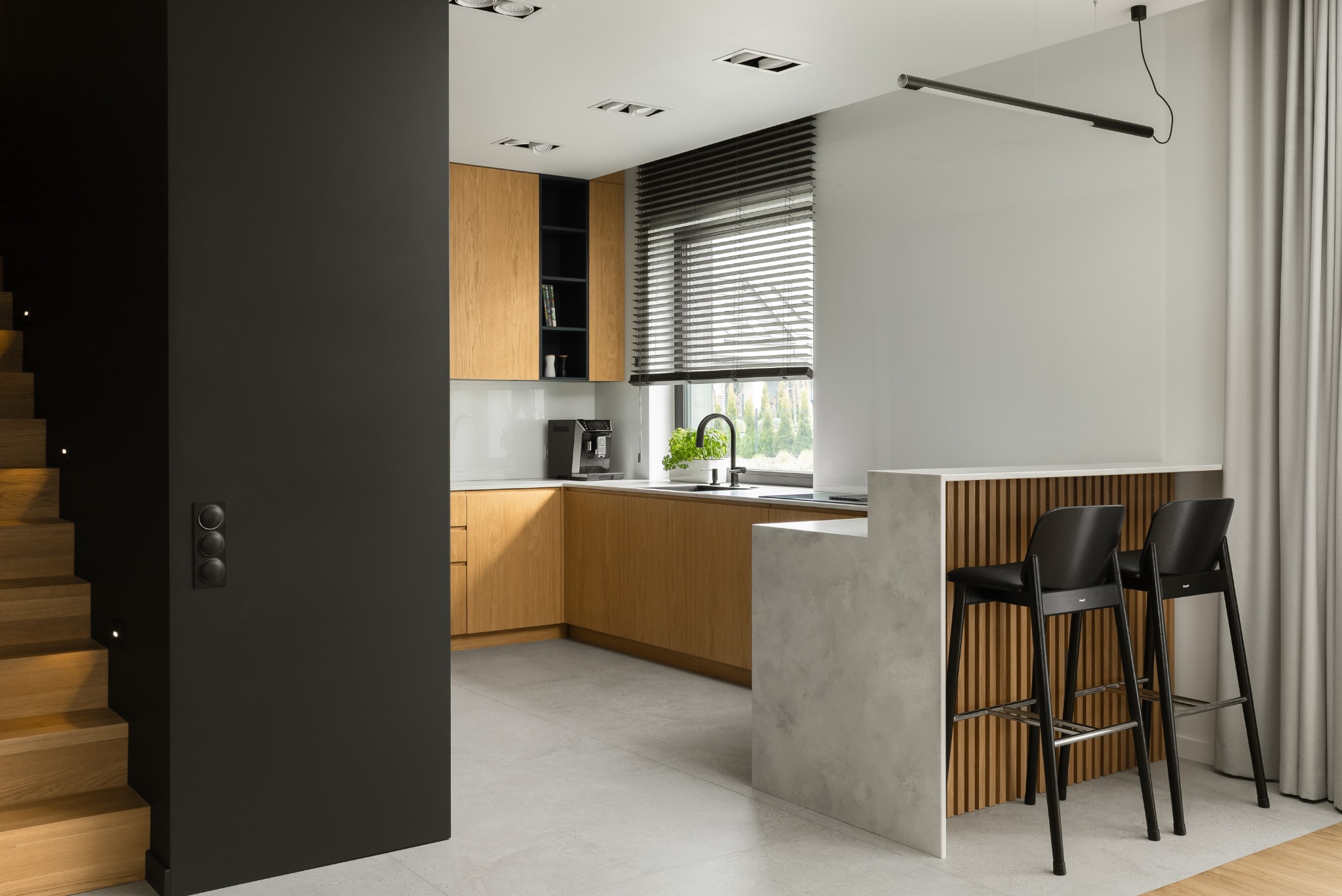 Nowoczesna kuchnia z drewnianymi szafkami, wyspą z hokerami i schodami z boku. Minimalistyczne oświetlenie i jasna kolorystyka. Widok z perspektywy wejścia.