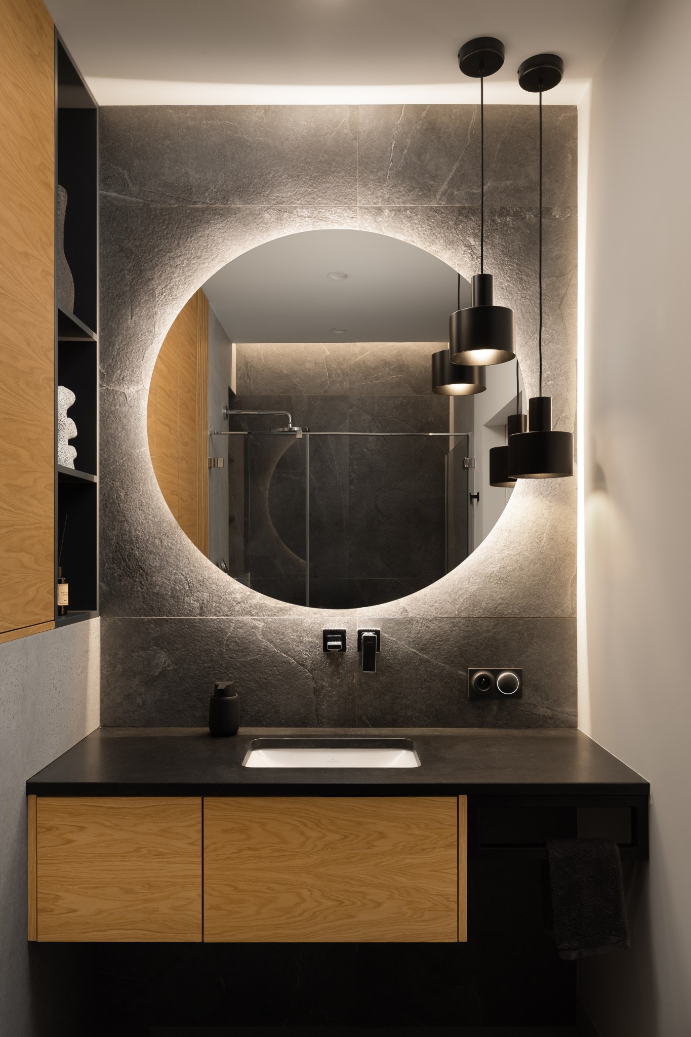 Nowoczesna łazienka w Pruszczu Gdańskim: okrągłe lustro z podświetleniem LED, czarny blat i drewniane szafki. Minimalistyczny design i eleganckie wykończenie.