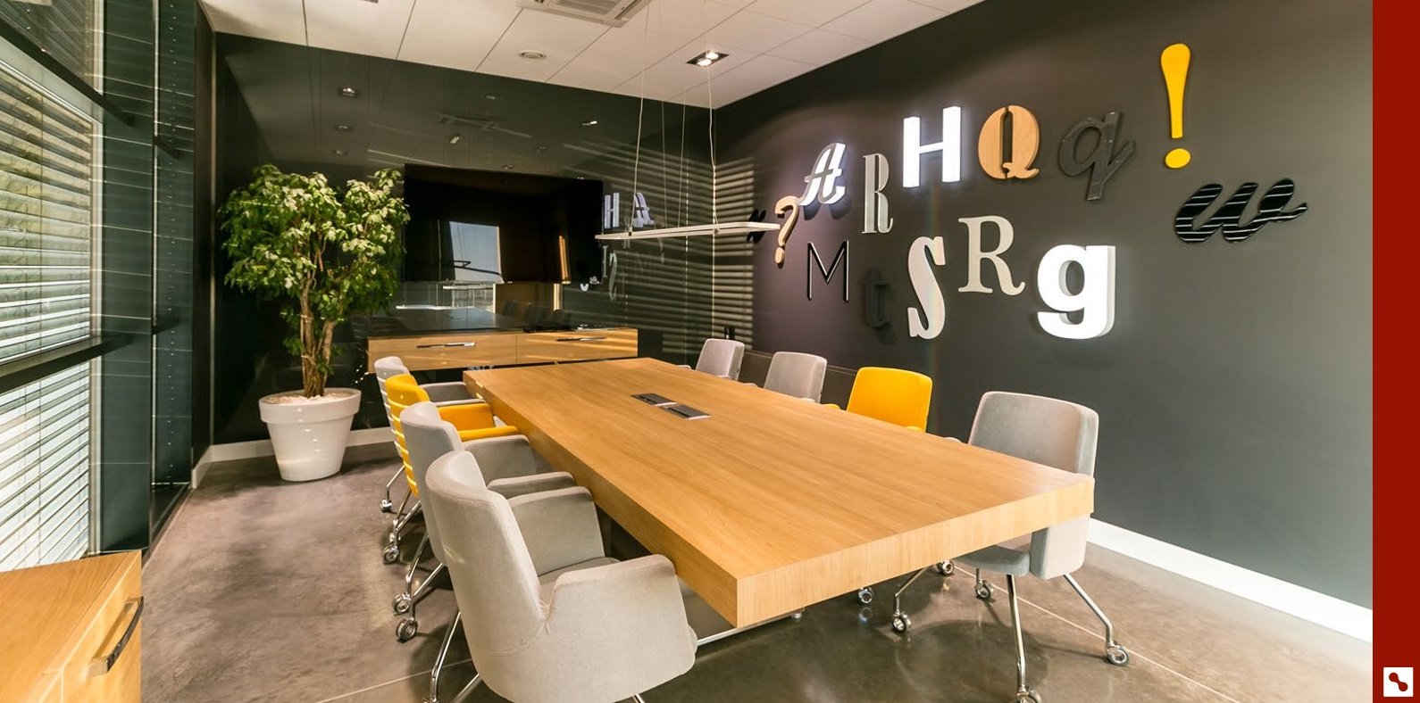 Sala konferencyjna z dużym drewnianym stołem, szarymi i żółtymi krzesłami na kółkach, ścianą z literami i telewizorem w tle.