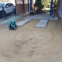 Przygotowanie podłoża z użyciem zagęszczarki wibracyjnej, widoczne płyty betonowe, piasek i samochód osobowy w tle.