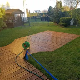 Świeżo wyczyszczony taras z drewna w ogrodzie, widoczny zwinięty wąż ogrodowy i fragment trawnika, w tle domek na rowery.