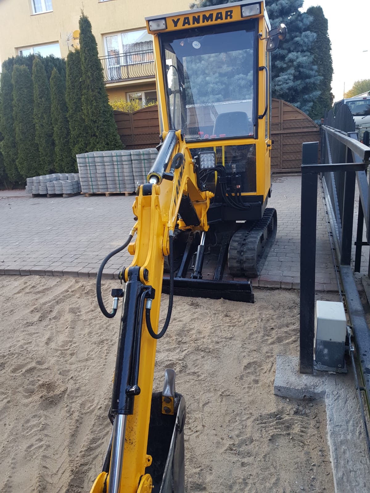 Żółta minikoparka Yanmar na piasku, obok kostka brukowa i brama wjazdowa.