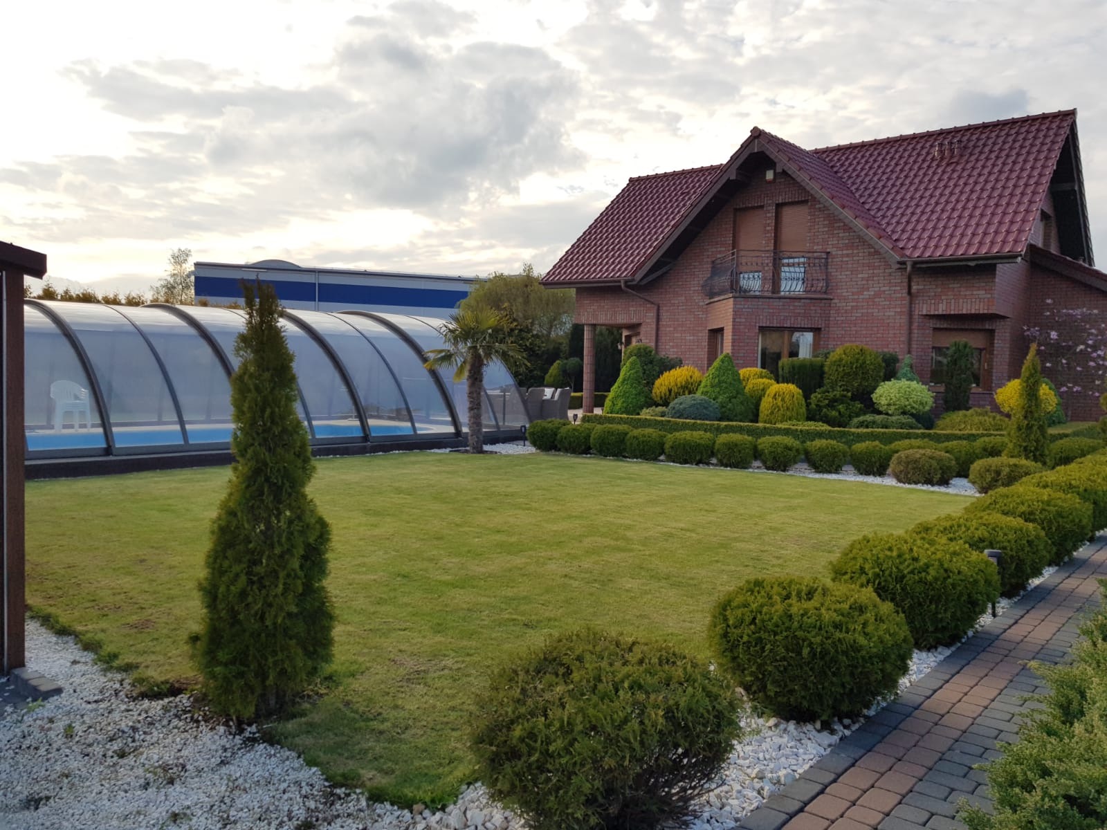 Zadbany ogród z równo przystrzyżonym trawnikiem, ozdobnymi krzewami i drzewami iglastymi, na tle domu z czerwonej cegły i zadaszonego basenu.