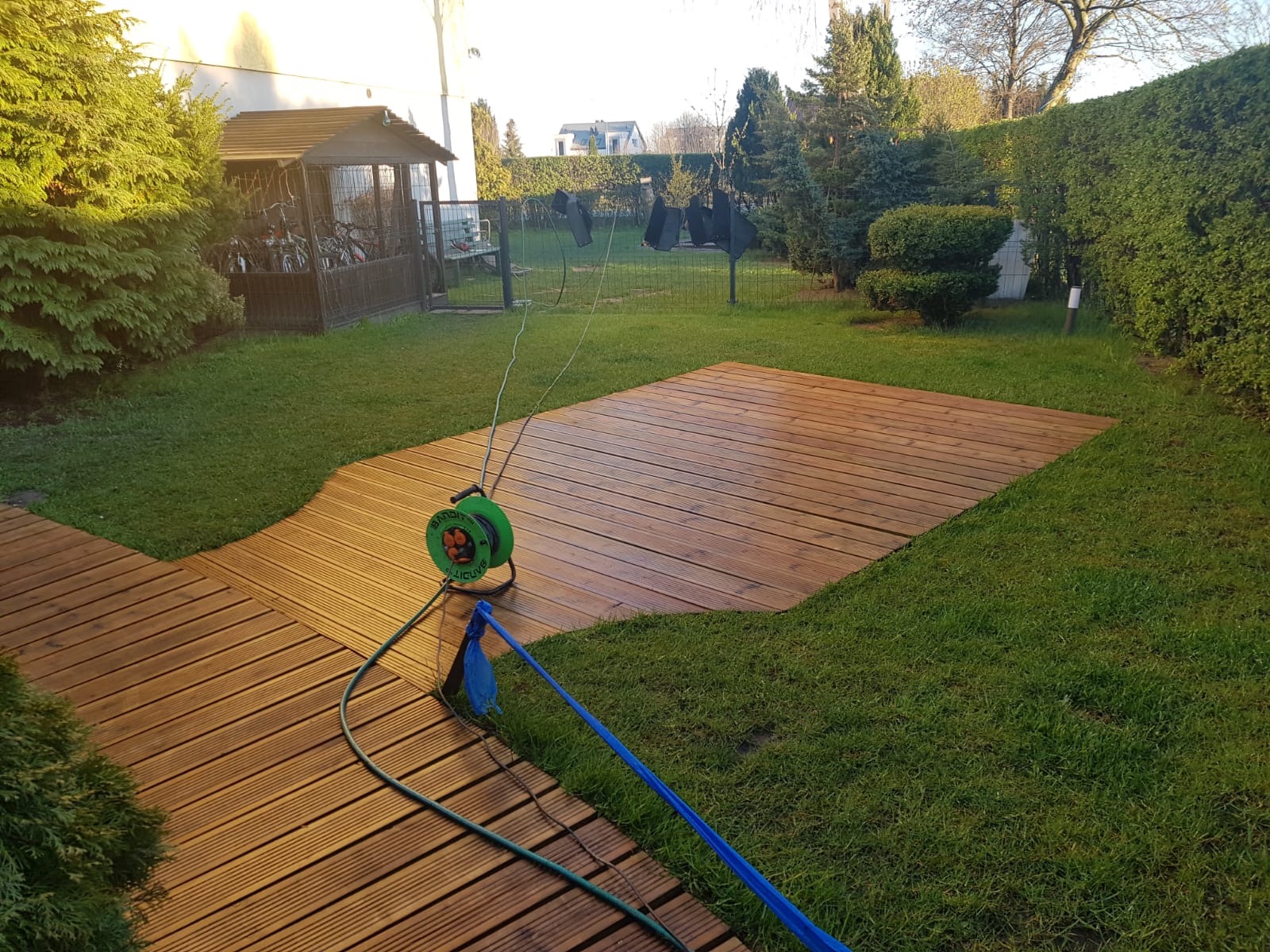 Świeżo wyczyszczony taras z drewna w ogrodzie, widoczny zwinięty wąż ogrodowy i fragment trawnika, w tle domek na rowery.